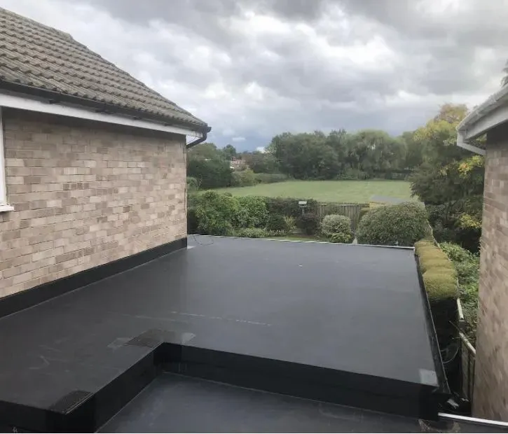 A completed flat roof installation on a home extension, showing the sleek, waterproof surface alongside the main tiled roof of the house and a distant field.