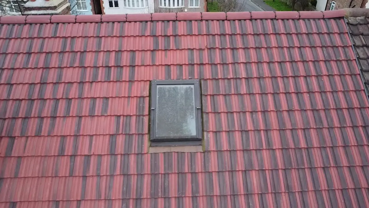 A close-up view of a tiled roof in shades of red and dark grey, with a rectangular Velux roof window installed in the centre.